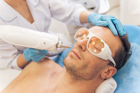 Handsome man is getting face skin treatment. Doctor in medical gloves is undertaking the procedure using a modern equipmentの写真素材