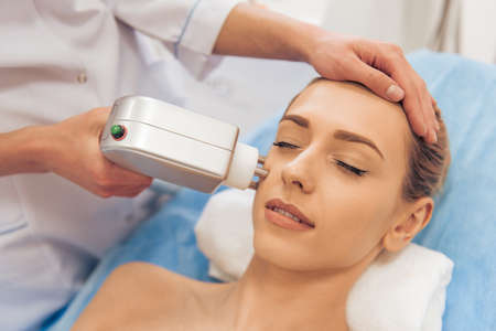 Beautiful woman is getting face skin treatment. Doctor is undertaking the procedure using a modern equipmentの写真素材