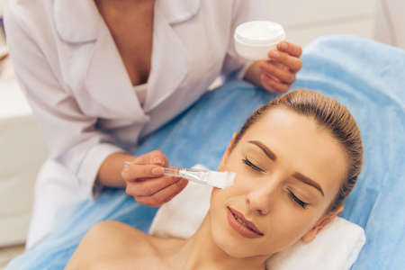 Beautiful young woman is getting face skin treatment. Cosmetician is holding a bottle of cream and using a brushの写真素材