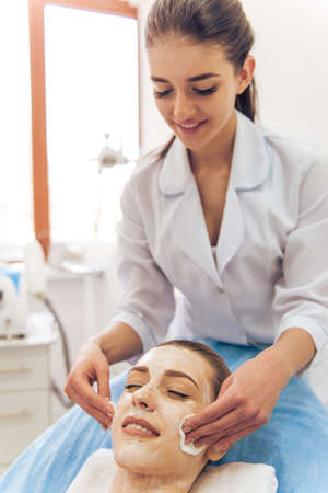 Beautiful young woman getting face skin treatment, lying with cream on her face. Female doctor is undertaking the procedureの写真素材