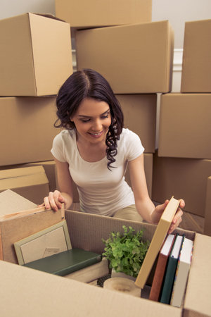 Attractive young woman is moving, smiling and looking through things while packing, sitting among...