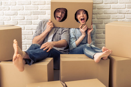 Happy young couple is moving, hiding heads in boxes with holes, looking at camera and making faces, sitting among other cardboard boxesの写真素材