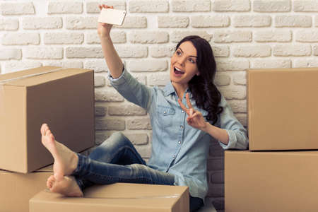 Attractive young woman is moving, making selfie using a smart phone and smiling while sitting among cardboard boxesの写真素材