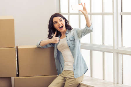 Attractive young woman is moving, standing among cardboard boxes, making selfie and smilingの写真素材