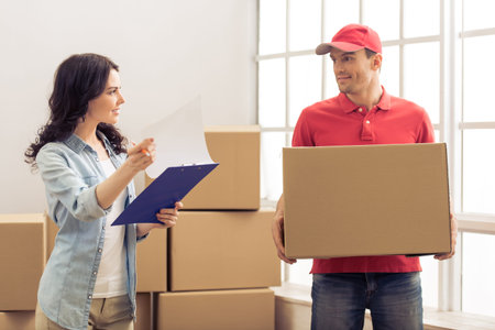 Attractive young woman is signing documents for moving. Handsome delivery worker is holding a...
