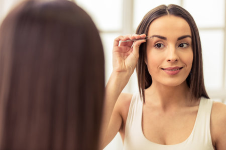 Beautiful young woman is plucking her eyebrows and smiling while looking at the mirrorの写真素材