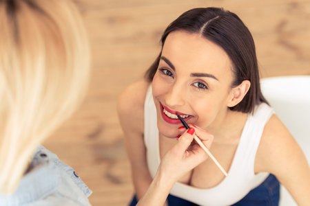 Top view of beautiful young woman looking at camera and smiling, visagiste is doing her makeup using a lip brushの写真素材