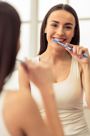 Beautiful young woman is brushing her teeth, looking at camera and smiling