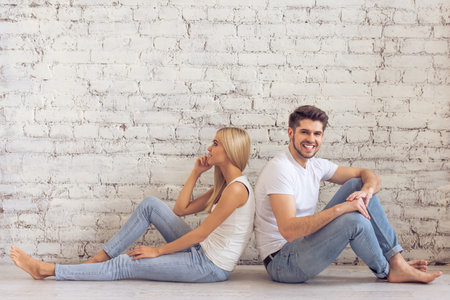 Beautiful young couple is sitting back to back on floor against white brick wall. Man is looking at camera and smilingの写真素材