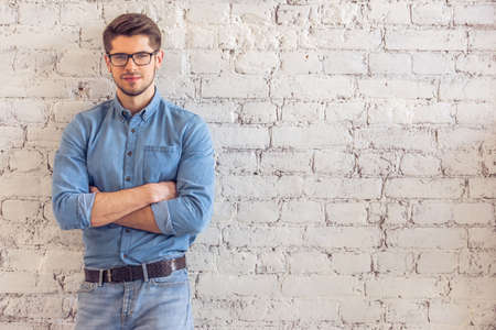 Portrait of handsome young man in jeans clothes and eyeglasses looking at camera and smiling, standing against white brick wallの写真素材