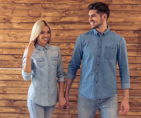 Beautiful young couple in jeans clothes is holding hands and smiling, standing against wooden wallの写真素材
