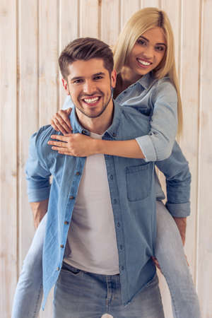 Beautiful young couple in jeans clothes is hugging, looking at camera and smiling, standing against wooden wall. Girl is pickabackの写真素材