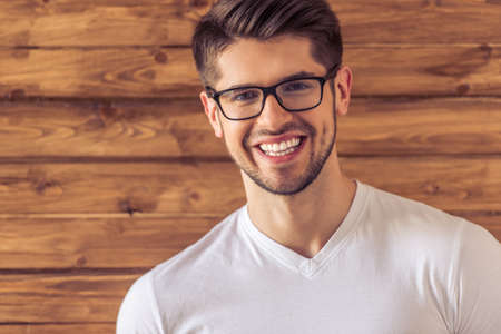 Portrait of handsome young man in eyeglasses looking at camera and smiling, against wooden wallの写真素材