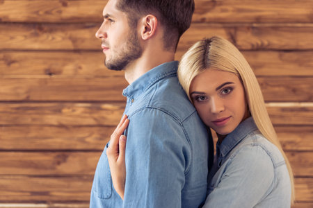 Portrait of beautiful young couple in jeans clothes standing against wooden wall. Girl is hugging her man and looking at cameraの写真素材