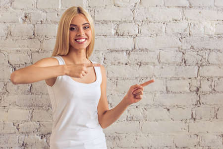 Attractive young woman in casual clothes is pointing, looking at camera and smiling, standing against white brick wallの写真素材