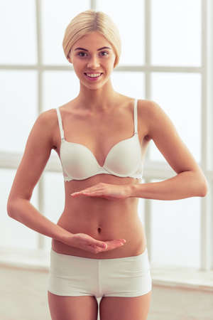 Beautiful blonde girl in white underwear is keeping hands in front of her belly, looking at camera and smilingの写真素材