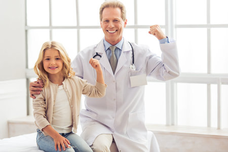 Cute little girl and handsome middle aged pediatrician are showing their biceps, looking at camera and smiling, sitting in doctors officeの写真素材