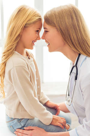 Beautiful young pediatrician and cute little girl are touching noses and smiling, sitting in doctors officeの写真素材
