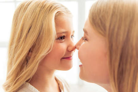 Beautiful young pediatrician and cute little girl are touching noses and smiling, sitting in doctors office, close-upの写真素材