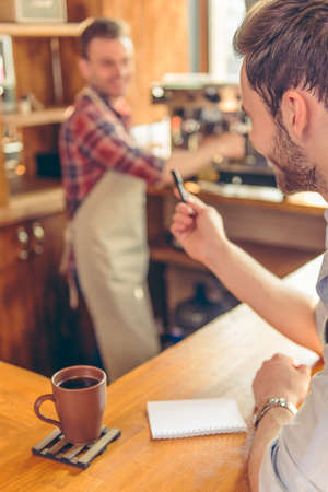Handsome young waiter is making notes and talking while sitting at the bar counter, in the background barista is making coffeeの写真素材