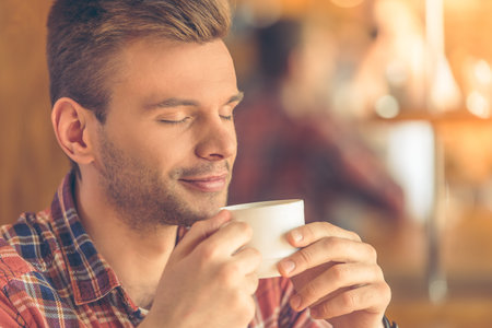 Portrait of handsome young man holding a cup and enjoying coffee while sitting in a modern urban cafeの写真素材