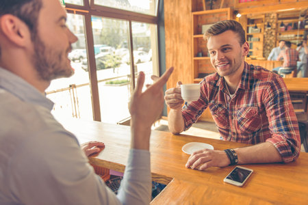 Handsome young man is drinking coffee, talking with barista and smiling, sitting at bar counter in a modern urban cafeの写真素材