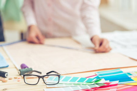 Glasses, thread and scissors lying among sketches and palette, in the background young designer is...