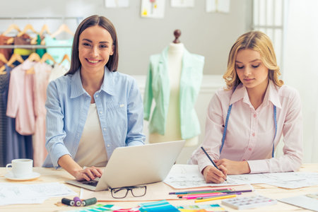 Beautiful young designers are working in dressmaking studio, using laptop and drawing sketches. One girl is looking at camera and smilingの写真素材