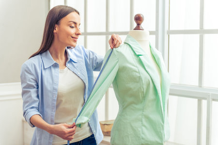 Beautiful young designer is smiling while working with mannequin and measure tape in dressmaking...