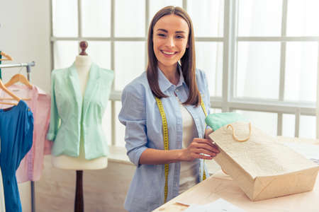 Beautiful young designer is holding a shopping bag, looking at camera and smiling while working in dressmaking studioの写真素材