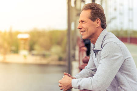 Side view of handsome middle aged man in sports uniform leaning on bridge and smiling while having break during morning runの写真素材