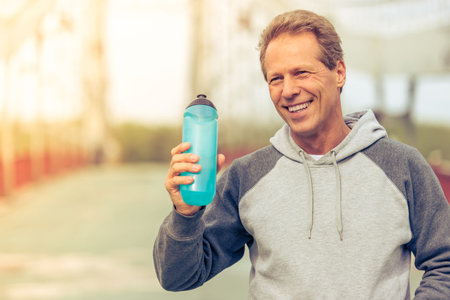 Handsome middle aged man in sports uniform is holding a bottle of water  and smiling during morning runの写真素材