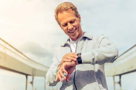 Handsome middle aged man in sports uniform and headphones is looking at his watch and smiling during morning runの写真素材