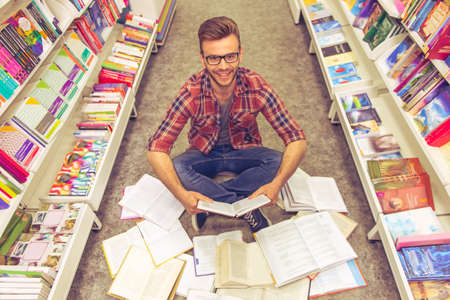 High angle view of handsome student reading a book, looking at camera and smiling while sitting on the floor among open books in the bookshopの写真素材