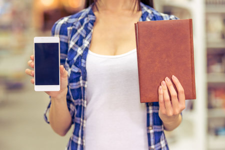 Cropped image of attractive girl holding a book and a smartphone while standing at the bookshop