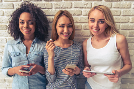 Beautiful young women of different nationalities are using gadgets, looking at camera and smiling, standing against white brick wallの写真素材