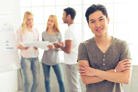 Handsome Asian guy is looking at camera and smiling, in the background his colleagues are discussing business affairsの写真素材