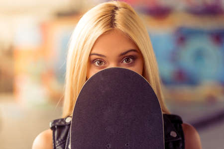 Portrait of beautiful blonde girl looking at camera and hiding face behind her skateboard while standing in skate parkの写真素材