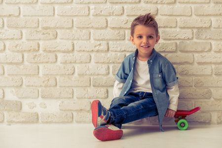 Cute stylish little boy in jeans is looking at camera and smiling, sitting on his skateboard against white brick wallの写真素材