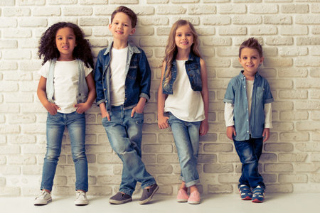 Full length portrait of cute little kids in stylish jeans clothes looking at camera and smiling, standing against white brick wallの写真素材