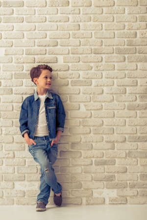 Full length portrait of stylish little boy in jeans clothes looking away and smiling, standing with hands in pockets against white brick wallの写真素材
