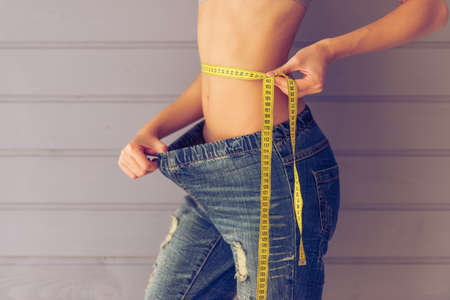 Cropped image of beautiful young slim woman pulling her jeans and using a tape measure showing loss of weight, on a gray backgroundの写真素材