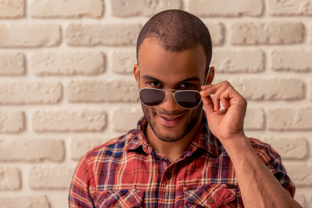 Portrait of handsome Afro American man in glasses looking at camera and smiling, against white brick wallの写真素材