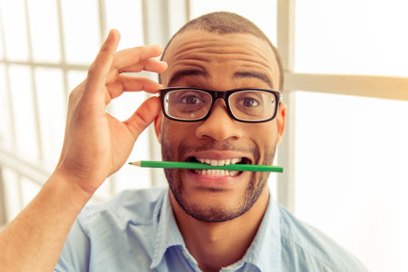 Portrait of handsome Afro American businessman in eyeglasses with pencil in his teeth looking at...