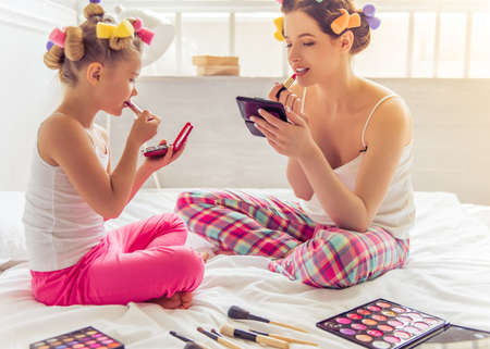 Beautiful young mother and her daughter in pajamas and with hair curlers are doing makeup, sitting on bed at homeの写真素材