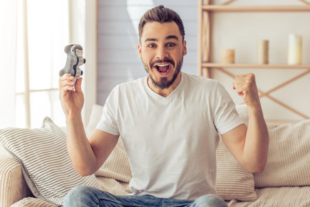 Handsome happy man is playing game console and showing success while sitting on the sofa at home