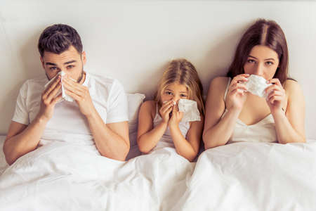 Beautiful young parents and their daughter are having common cold, wiping noses and looking at camera while lying on bedの写真素材