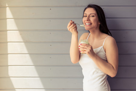 Attractive young woman in underwear is eating yogurt, looking at camera and smiling, standing against grey wallの写真素材