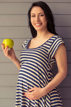 Beautiful pregnant woman is holding an apple, keeping one hand on a belly, looking at camera and smilingの写真素材
