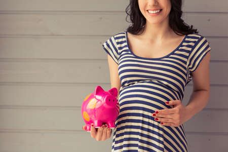 Cropped image of beautiful pregnant woman holding a piggy bank, keeping one hand on a belly and smiling, standing against gray wallの写真素材